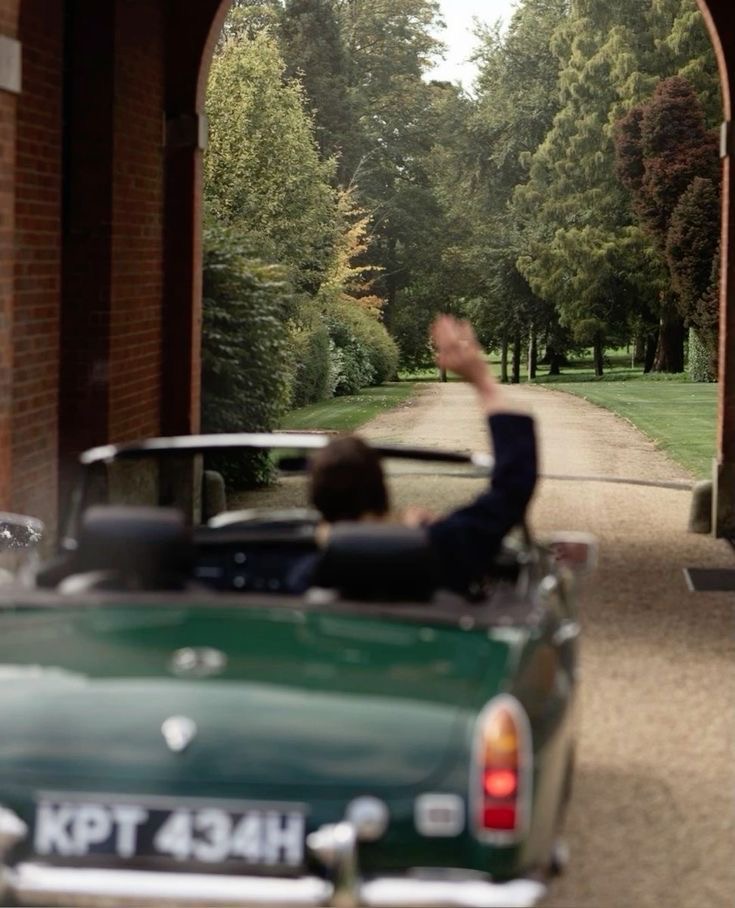 A vintage convertible driving through an arched estate entrance lined with trees