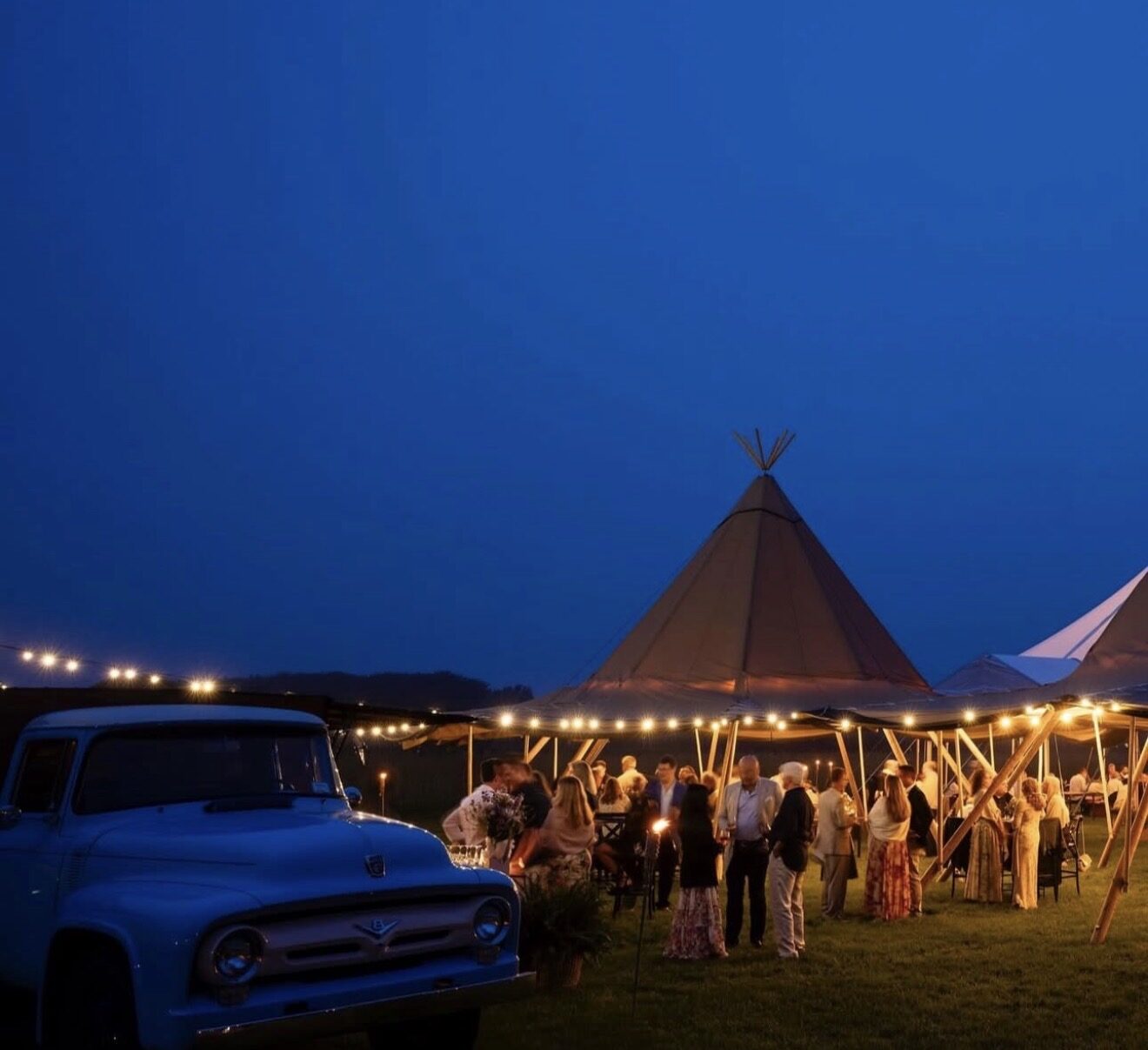 Guests under string lights and an open tent at the farm at blue hour